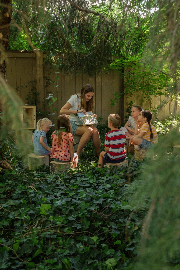 Teacher reading a book to children sitting on logs in a garden
